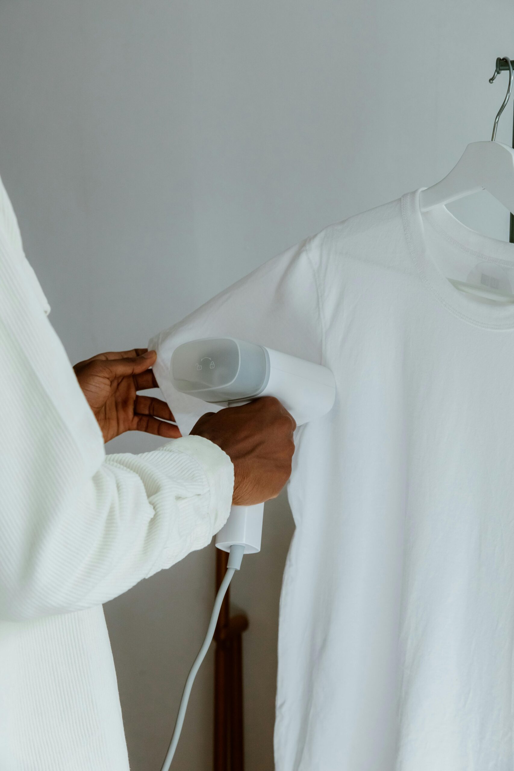 Hands using a steam iron on a white shirt hanging indoors.